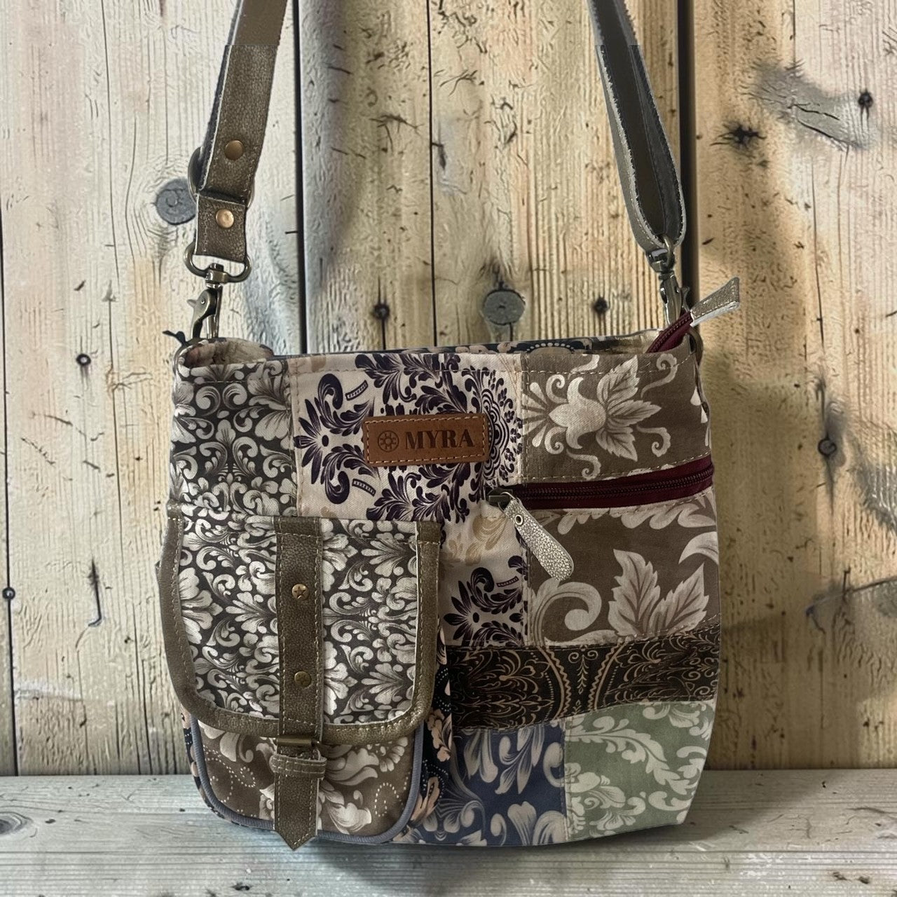 A patchwork fabric shoulder bag with floral and paisley patterns in beige, brown, navy, and green, featuring a brown leather brand patch that reads 'MYRA'.