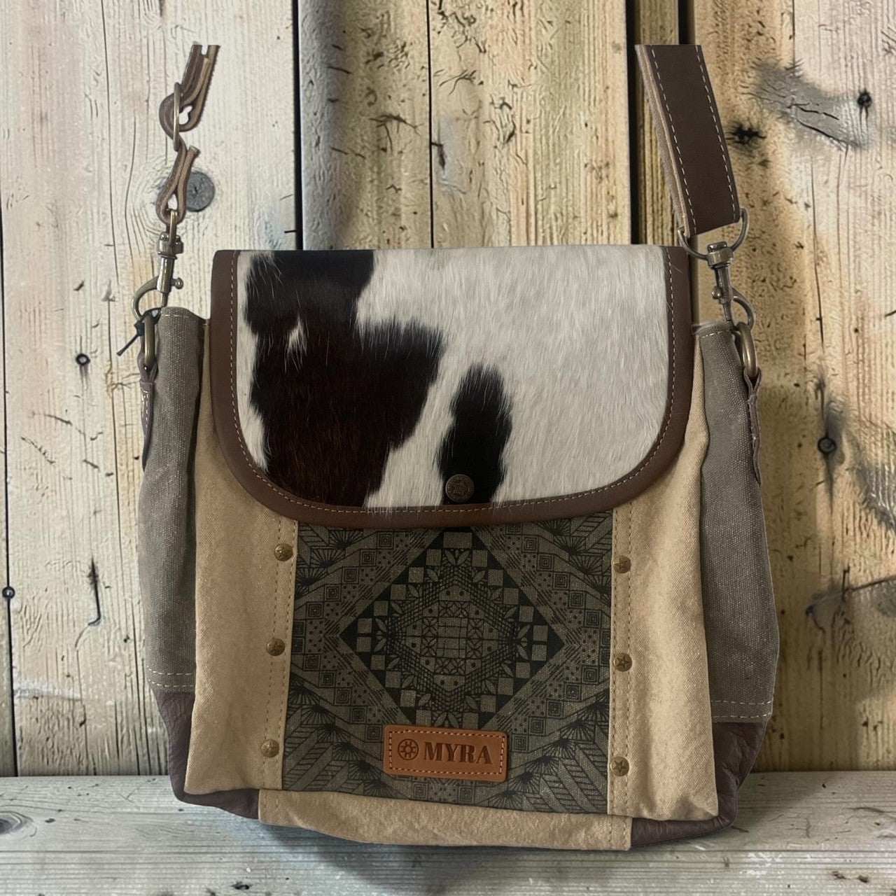 A western-style shoulder bag with a cowhide flap, canvas body, and brown leather trim, displayed against a rustic wooden plank background. The bag features a geometric patterned panel and a small 'MYHA' leather patch.