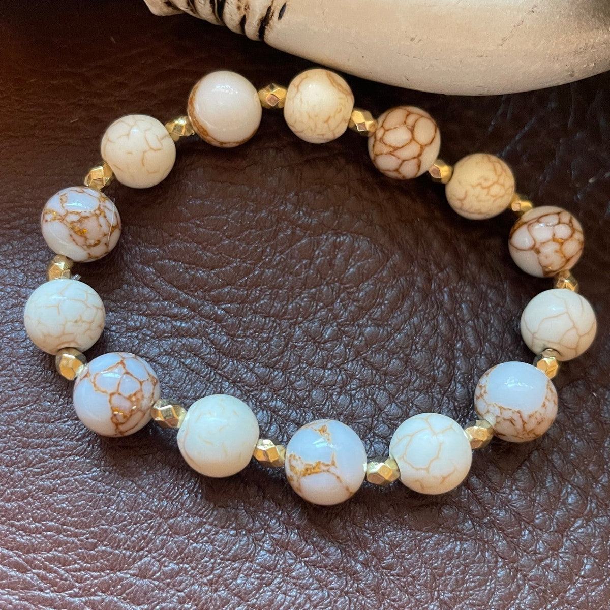 A beaded stretch bracelet featuring round white and cream marbled beads with brown veining, separated by small gold spacer beads, displayed on a brown textured leather surface.