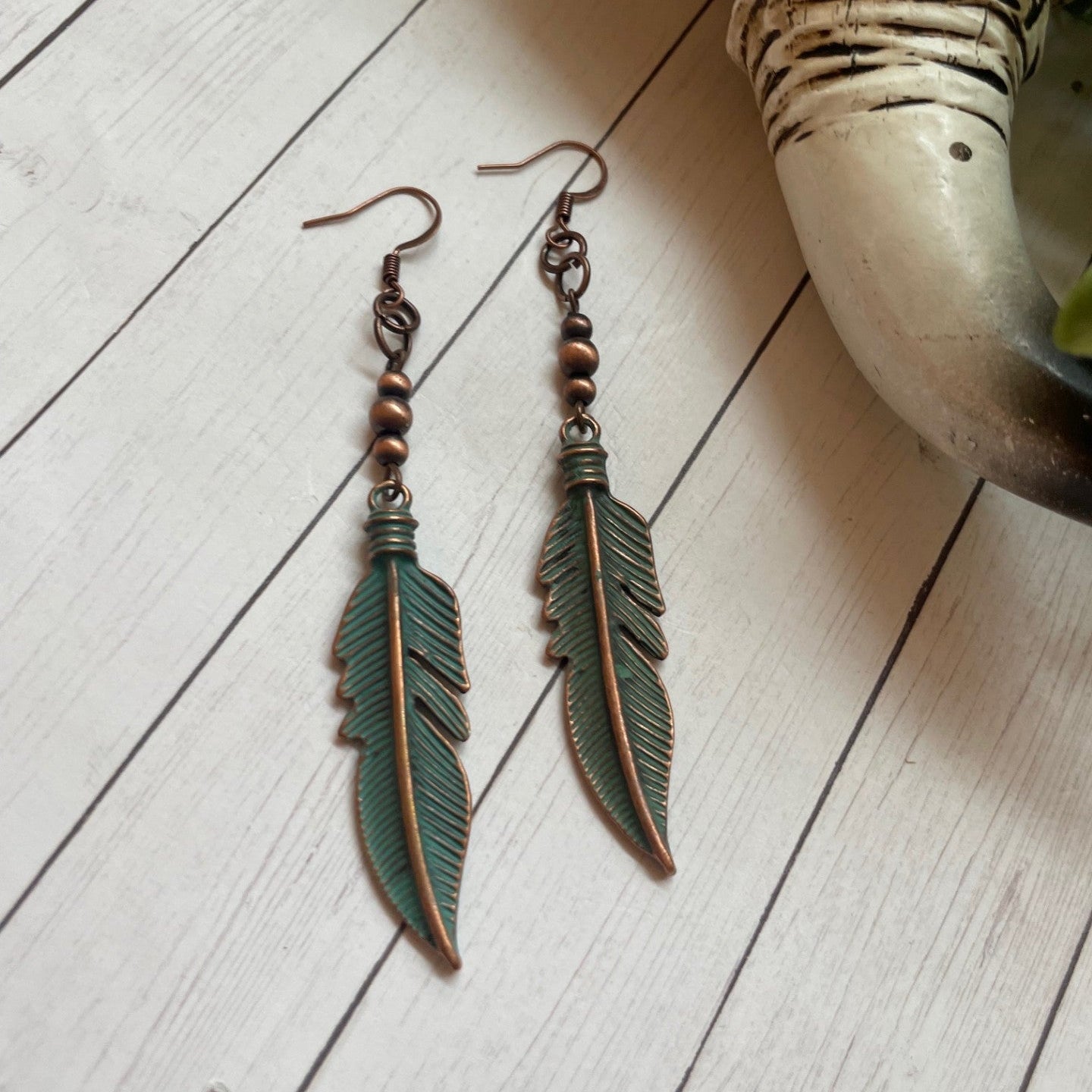 Feather-shaped earrings on a light wooden surface with a plant pot in the background.