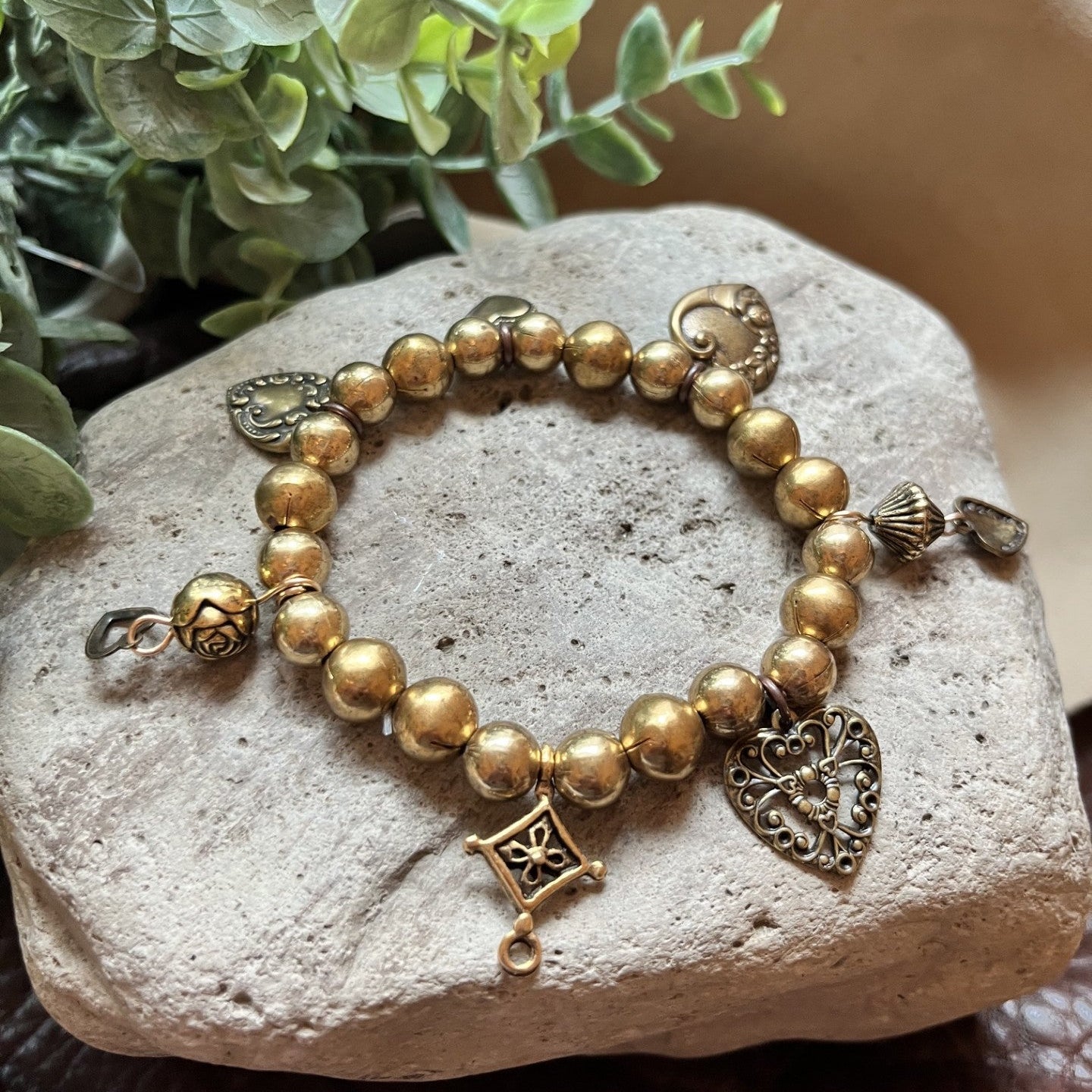A beaded bracelet with metallic gold-toned round beads, filigree heart charm, and decorative metal accents, displayed on a light stone surface with green leaves and a beige fabric background.