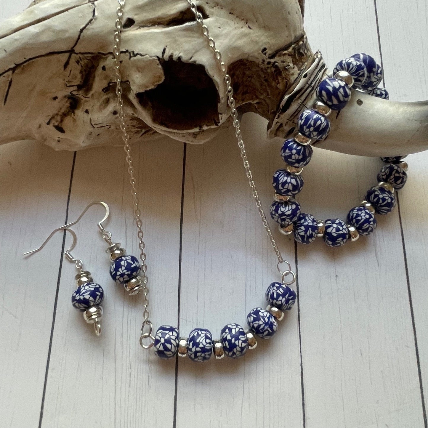 A jewelry set featuring blue and white patterned beads on a silver chain, displayed next to a bone skull on a white wood plank background. The set includes a bracelet, a necklace, and matching drop earrings with silver-tone fishhook wires.