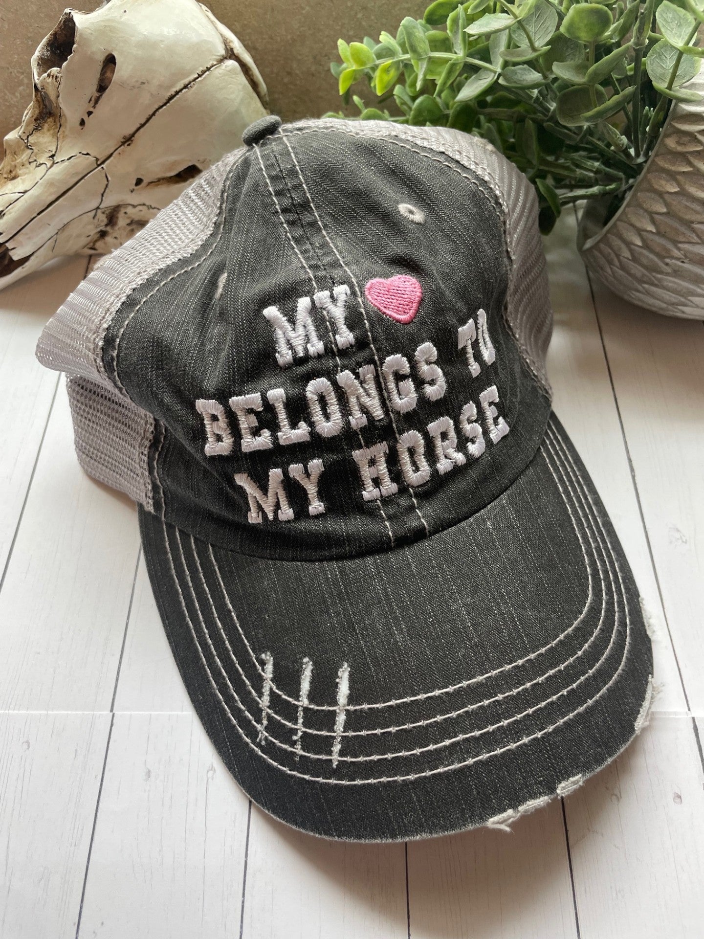 Black distressed cap with 'My Heart Belongs to My Horse' text on a wooden surface with a plant in the background.