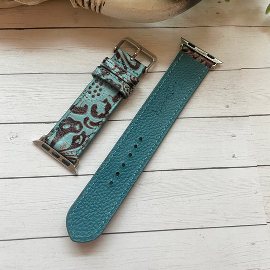 Two teal patterned watch straps on a wooden surface with a plant in the corner.