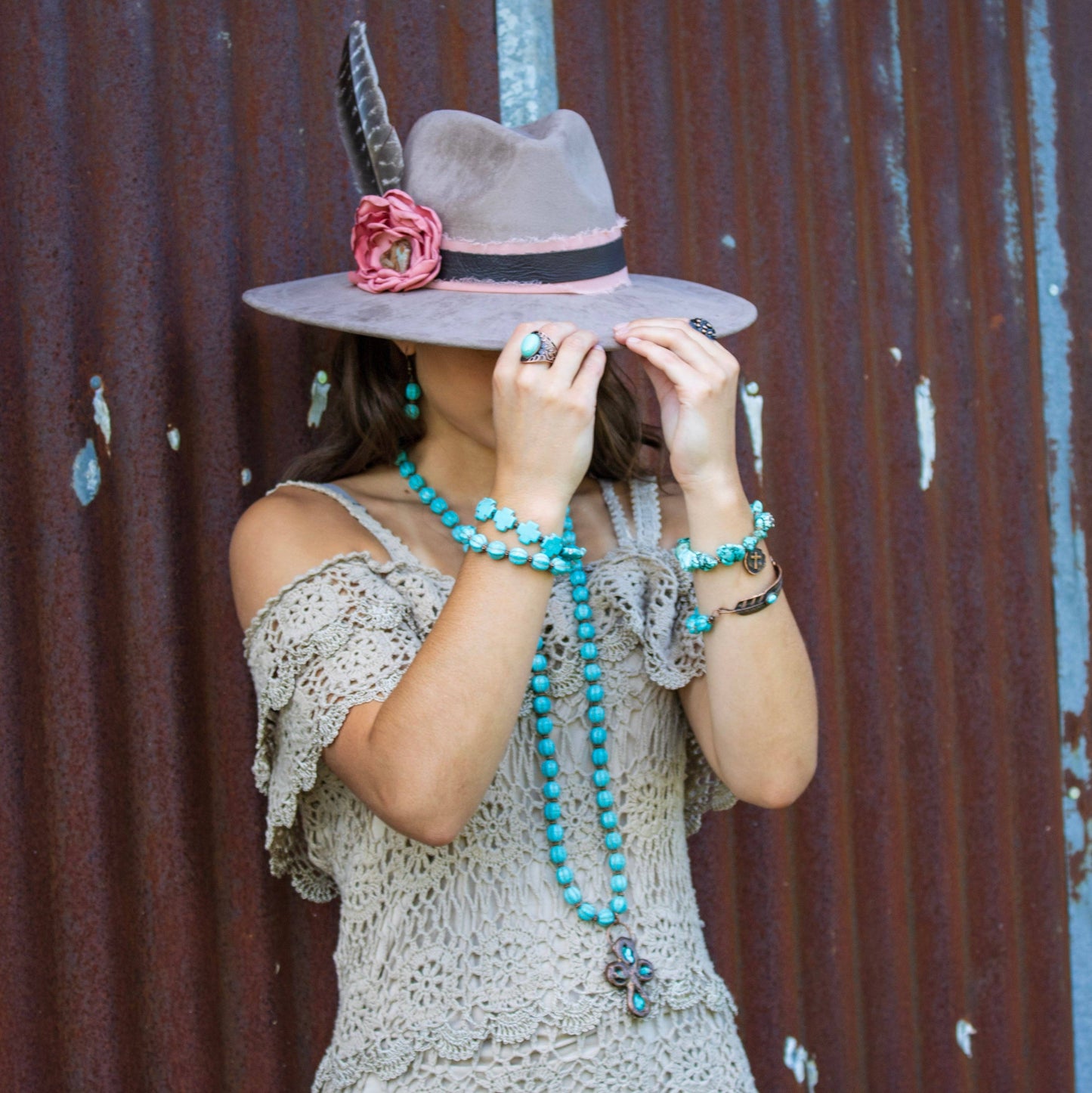 Person wearing a wide-brimmed hat with a feather and flower, standing against a rusty metal wall.