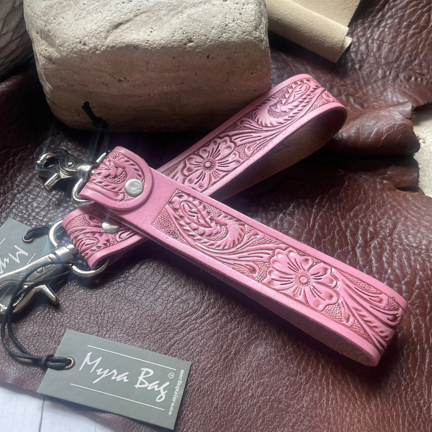 A pink tooled leather keychain or bag charm with floral embossing, shown with metal hardware and a branded paper tag reading Myra Bag, placed on a brown leather surface with stones in the background.