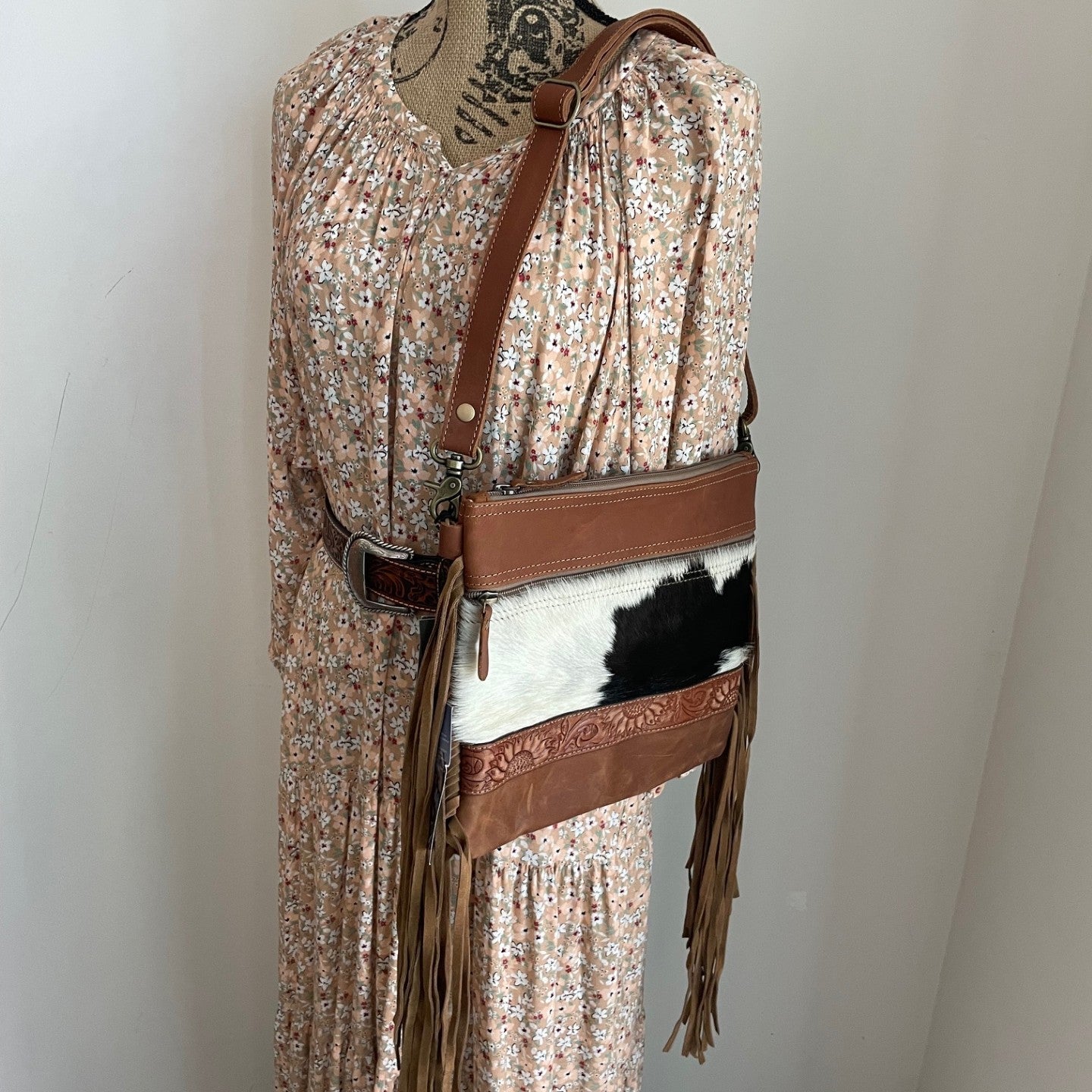 Mannequin wearing a floral dress with a brown leather belt and cowhide pouch against a white wall.