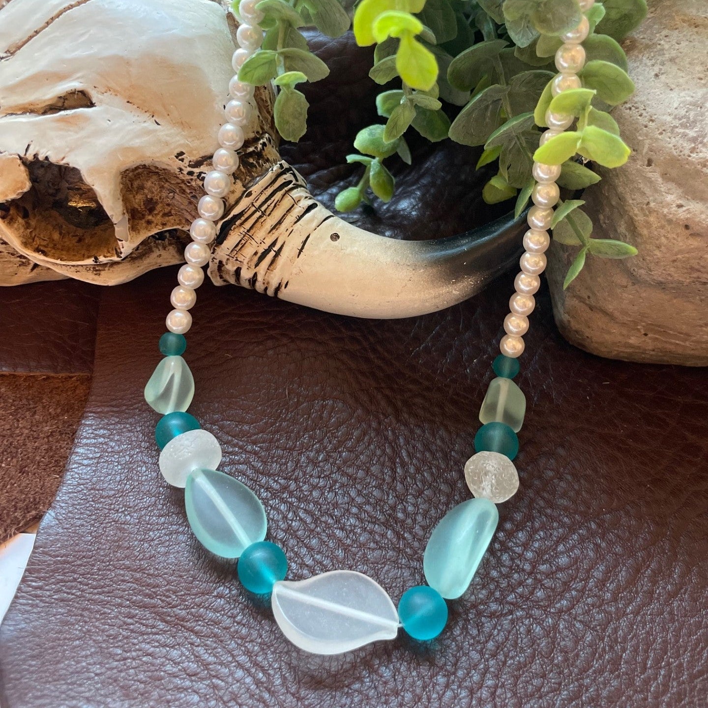 A beaded necklace with large translucent light blue and white teardrop beads, smaller turquoise blue beads, and strands of white pearl-like beads, displayed on a brown leather surface with decorative shells and greenery in the background.