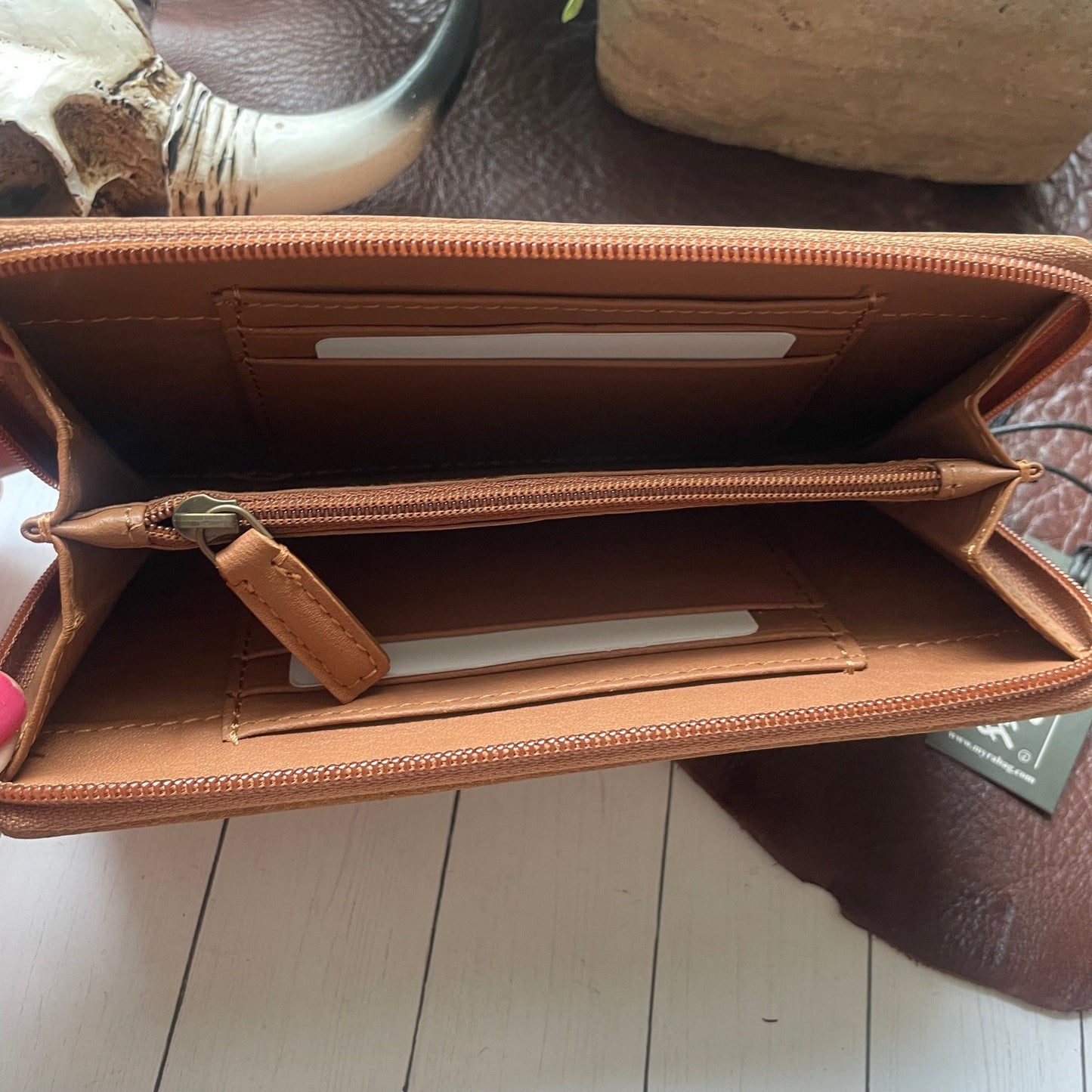 An open tan leather zip-around wallet or money clip shown from the inside, with card slots, a zip compartment, and interior pockets visible on a light wood floor.