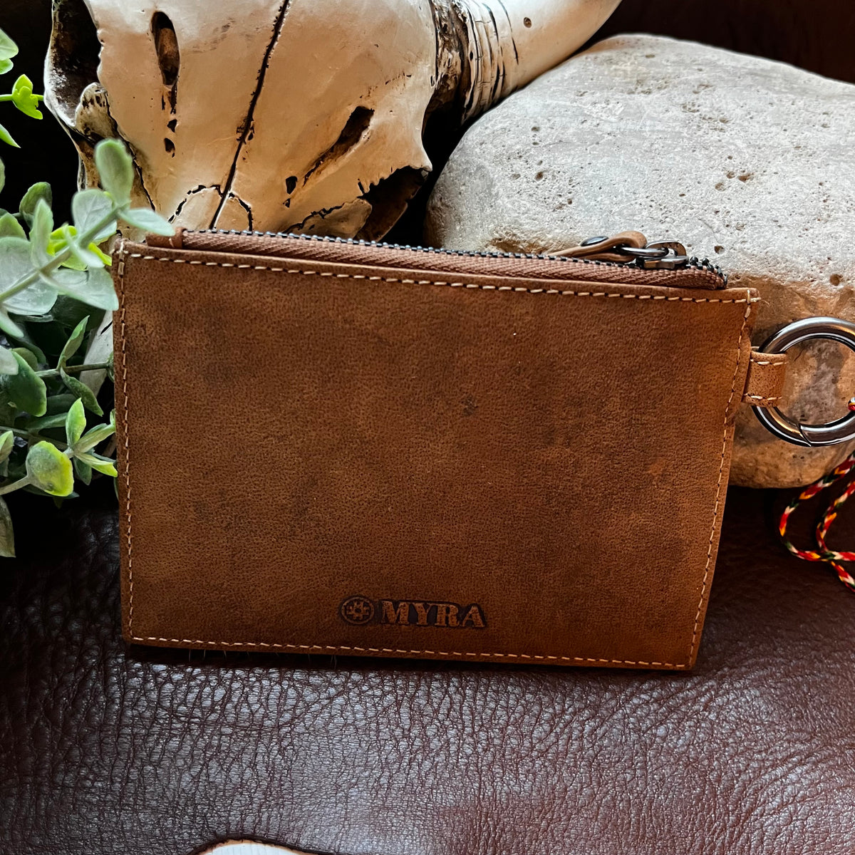 A brown leather zippered pouch with a metal ring attachment, featuring the debossed brand name 'MYRA' on the front, styled on a dark leather surface with a skull and greenery in the background.