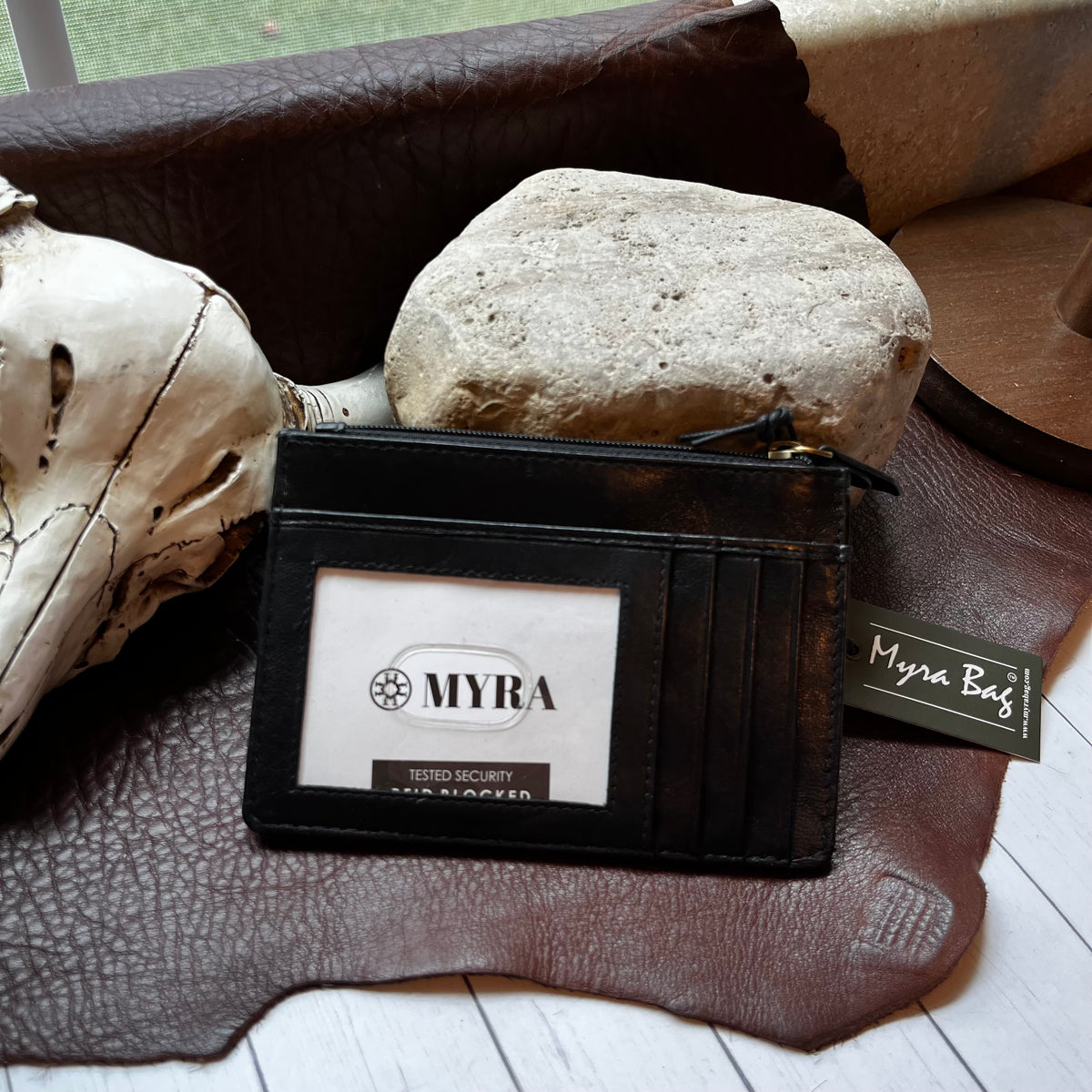 A black leather card holder wallet with a clear ID window displaying the brand name 'MYRA' and a small 'Myra Bag' hangtag, styled on a brown leather hide with a skull and stone.