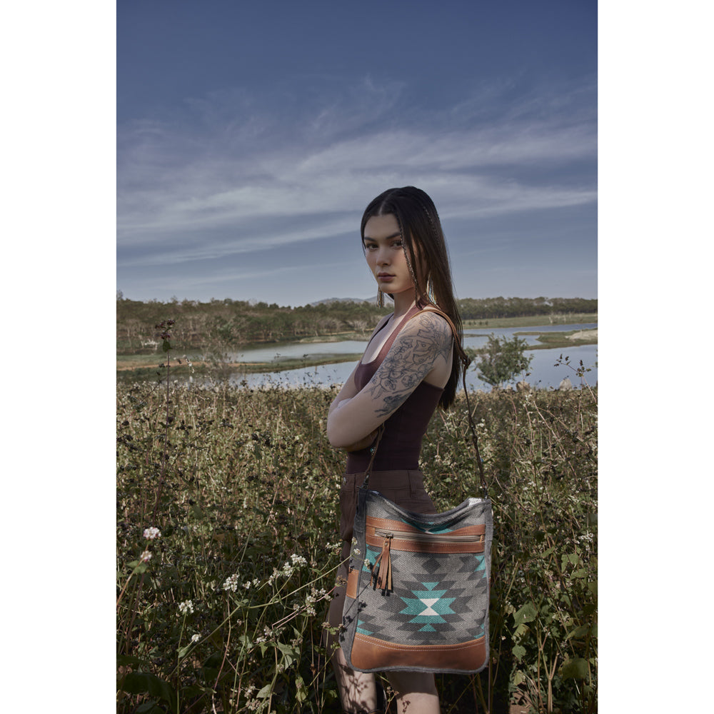 Woman holding a patterned bag in a natural setting with water and sky.