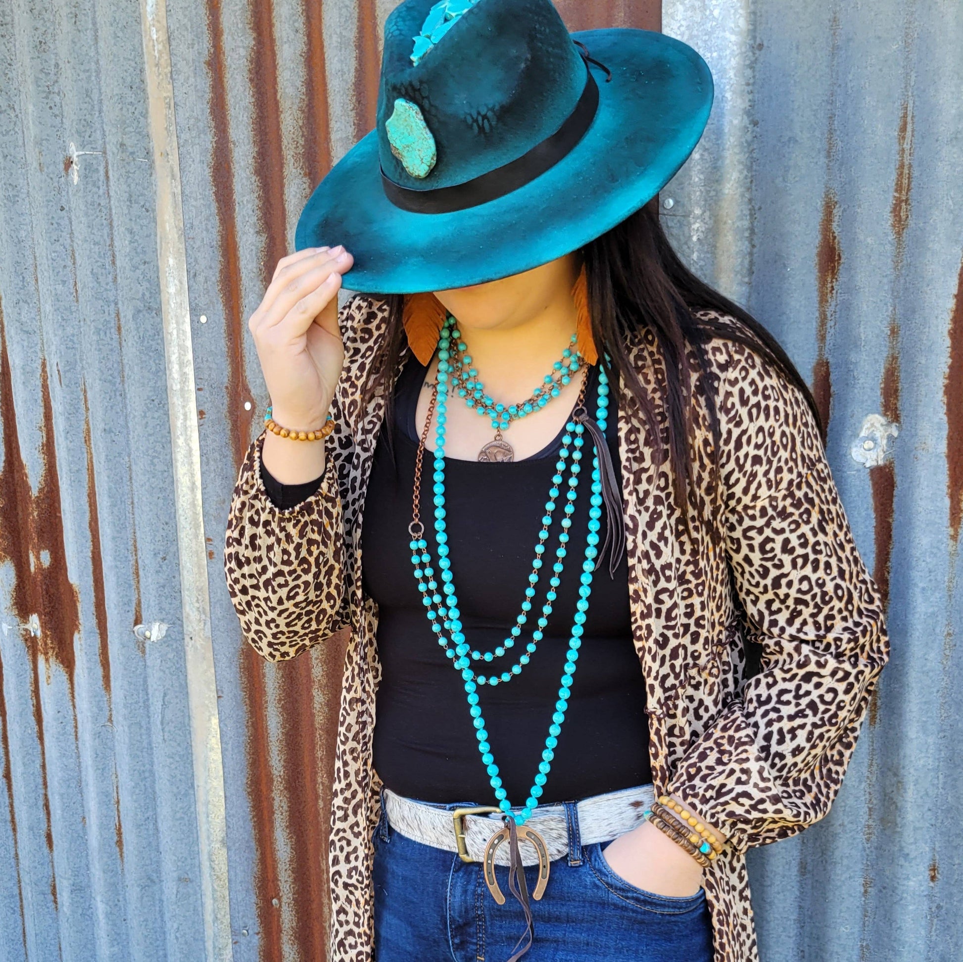 Person wearing a leopard print cardigan, black top, blue jeans, and turquoise hat and jewelry against a rustic metal wall.