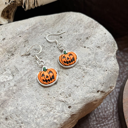 A pair of silver dangle earrings shaped like orange jack-o'-lantern pumpkins with carved black faces and small green stems, displayed on a textured gray stone.
