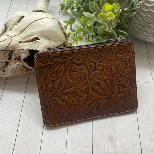 Brown leather wallet with floral embossing on a light wooden surface with a skull and plant in the background.