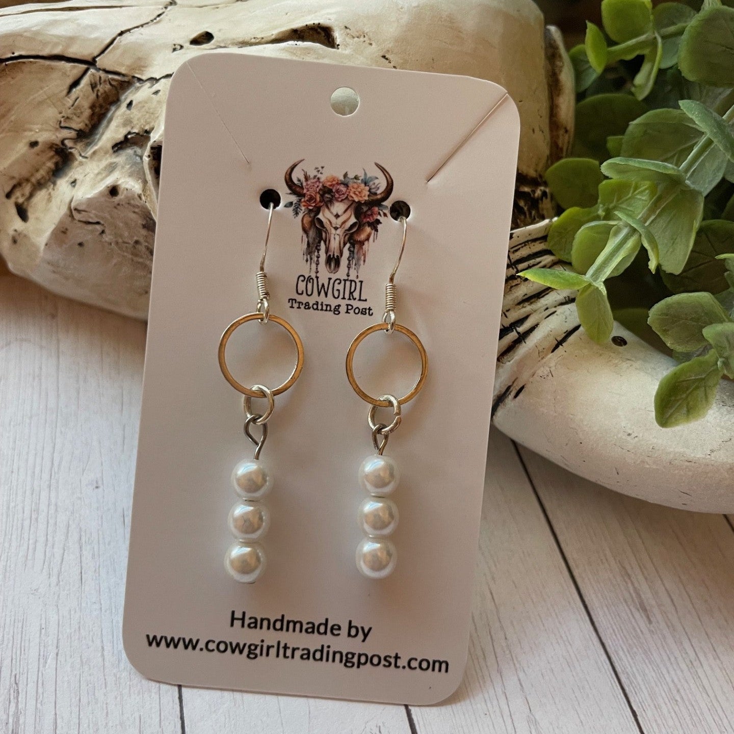 Pair of gold and pearl earrings on a 'Cowgirl Trading Post' card with a rustic background.