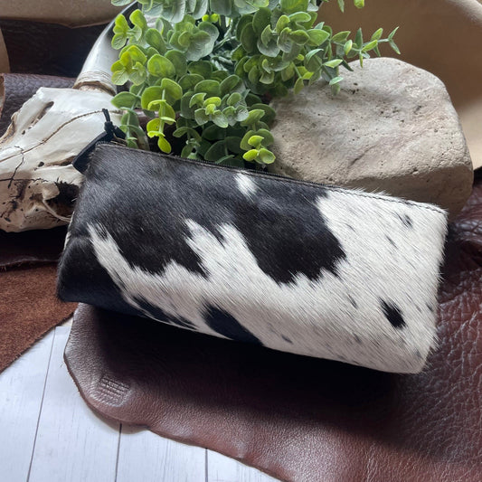 A black and white cowhide zippered pouch resting on a brown leather surface with a green plant and stones in the background.