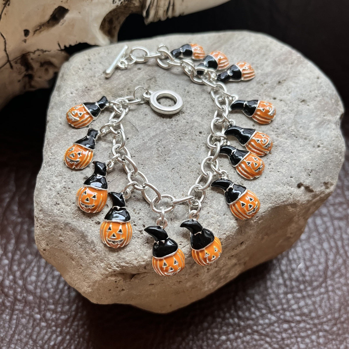 A silver charm bracelet with multiple small pumpkin and witch motifs, displayed on a textured gray stone over a dark brown surface.