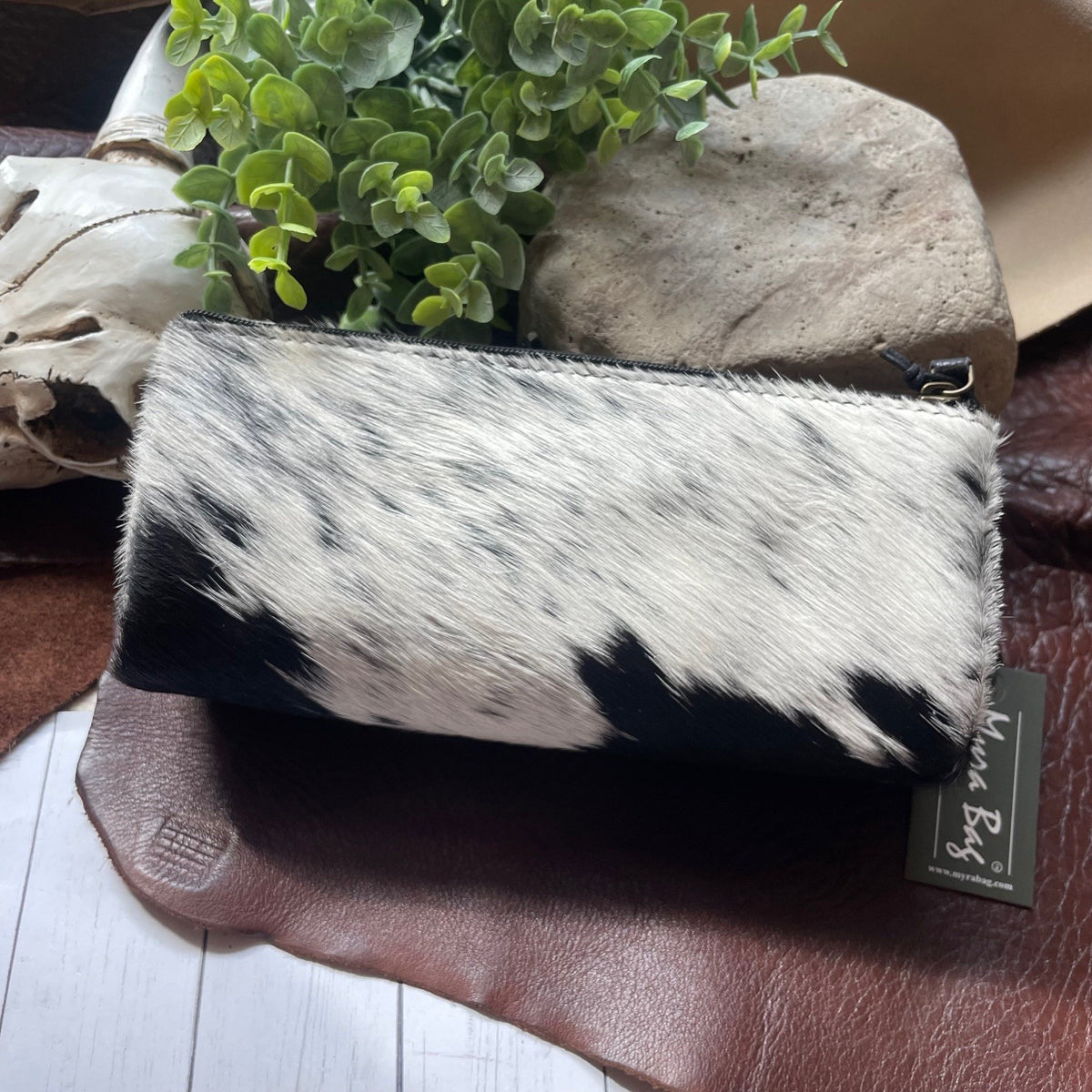 A cowhide zip-around pouch with a black and white mottled pattern, resting on a brown leather surface next to a small green plant and a stone.