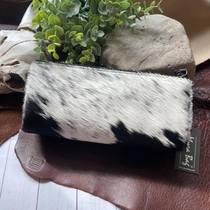 A cowhide zip-around pouch with a black and white mottled pattern, resting on a brown leather surface next to a small green plant and a stone.