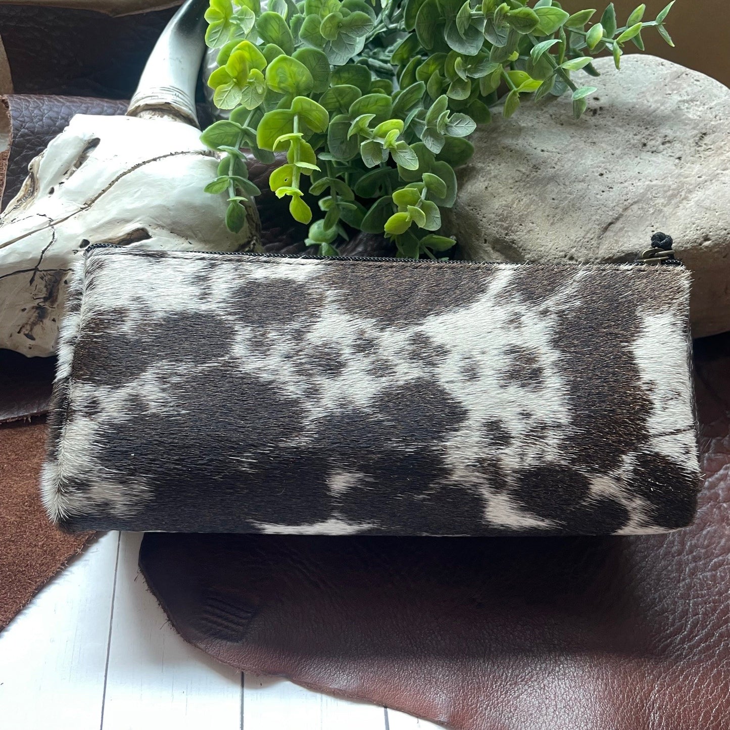 A rectangular zippered pouch with a black and white cowhide pattern, styled on a brown leather surface with a green plant and decorative stones in the background.
