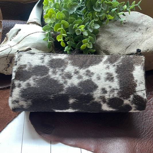 A rectangular zippered pouch with a black and white cowhide pattern, styled on a brown leather surface with a green plant and decorative stones in the background.