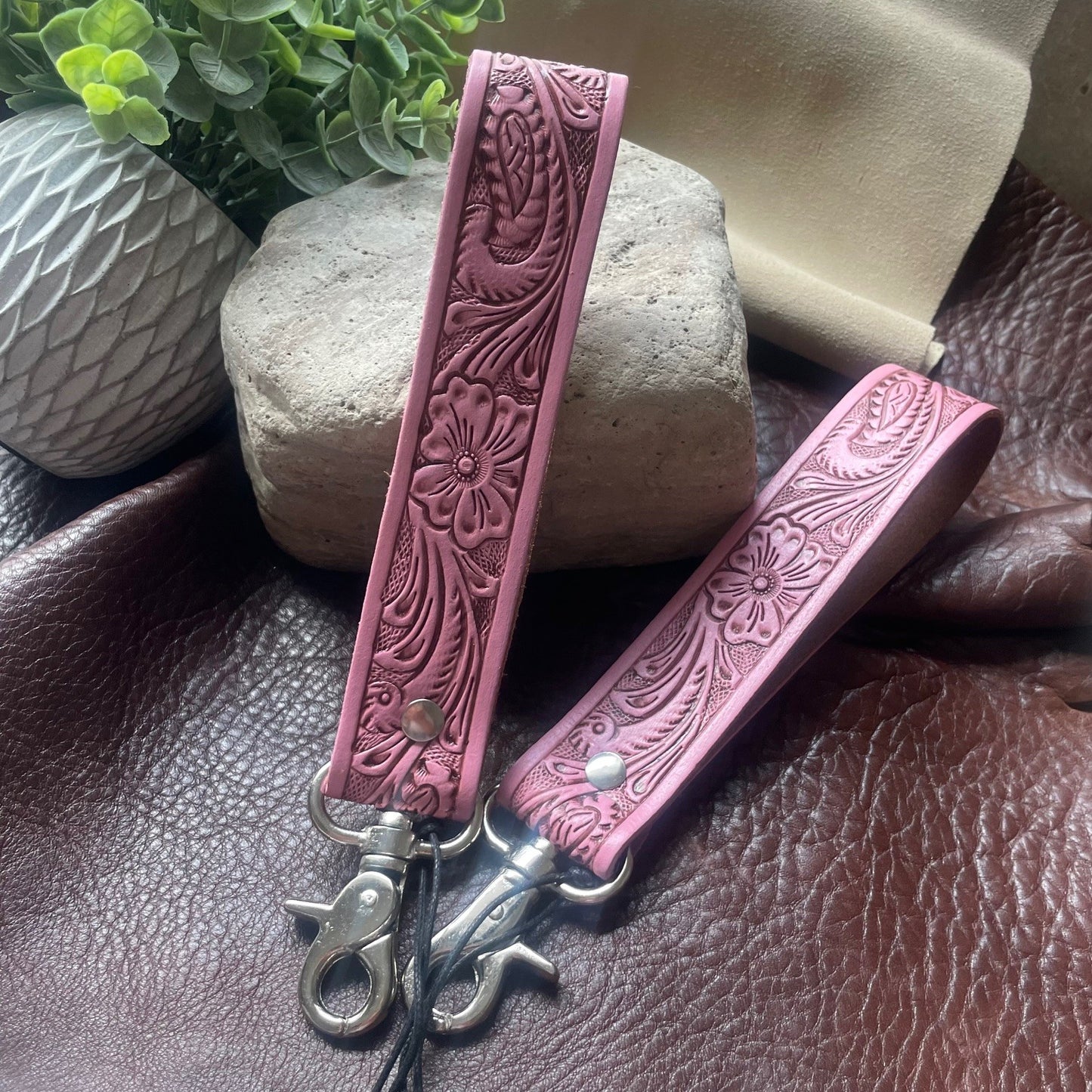 Two pink tooled leather wristlet keychains with floral embossing and silver-tone metal clasps, displayed on a brown leather surface with decorative stones and a potted plant in the background.
