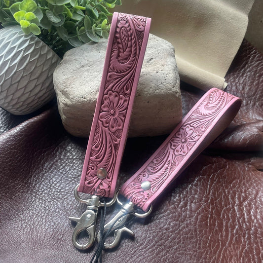 Two pink tooled leather wristlet keychains with floral embossing and silver-tone metal clasps, displayed on a brown leather surface with decorative stones and a potted plant in the background.