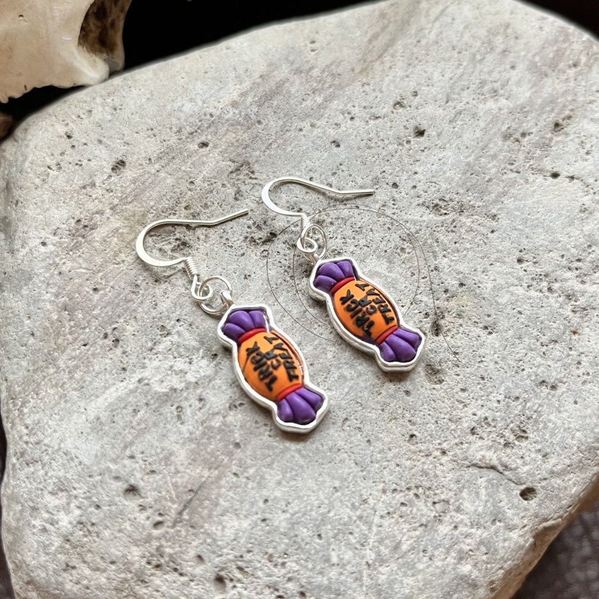 A pair of dangling earrings shaped like wrapped candies, with silver-tone hooks and bodies in orange and purple, resting on a light gray stone.