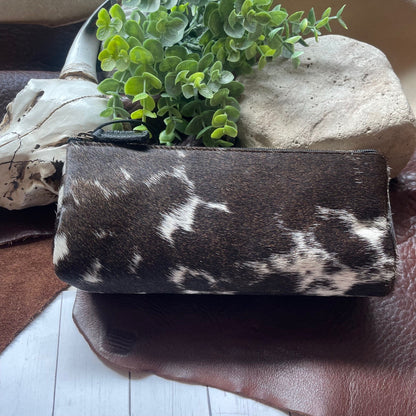 A zippered pouch or clutch in dark brown and white cowhide-patterned material, styled on a brown leather surface with a small green plant, a stone, and a skull prop.