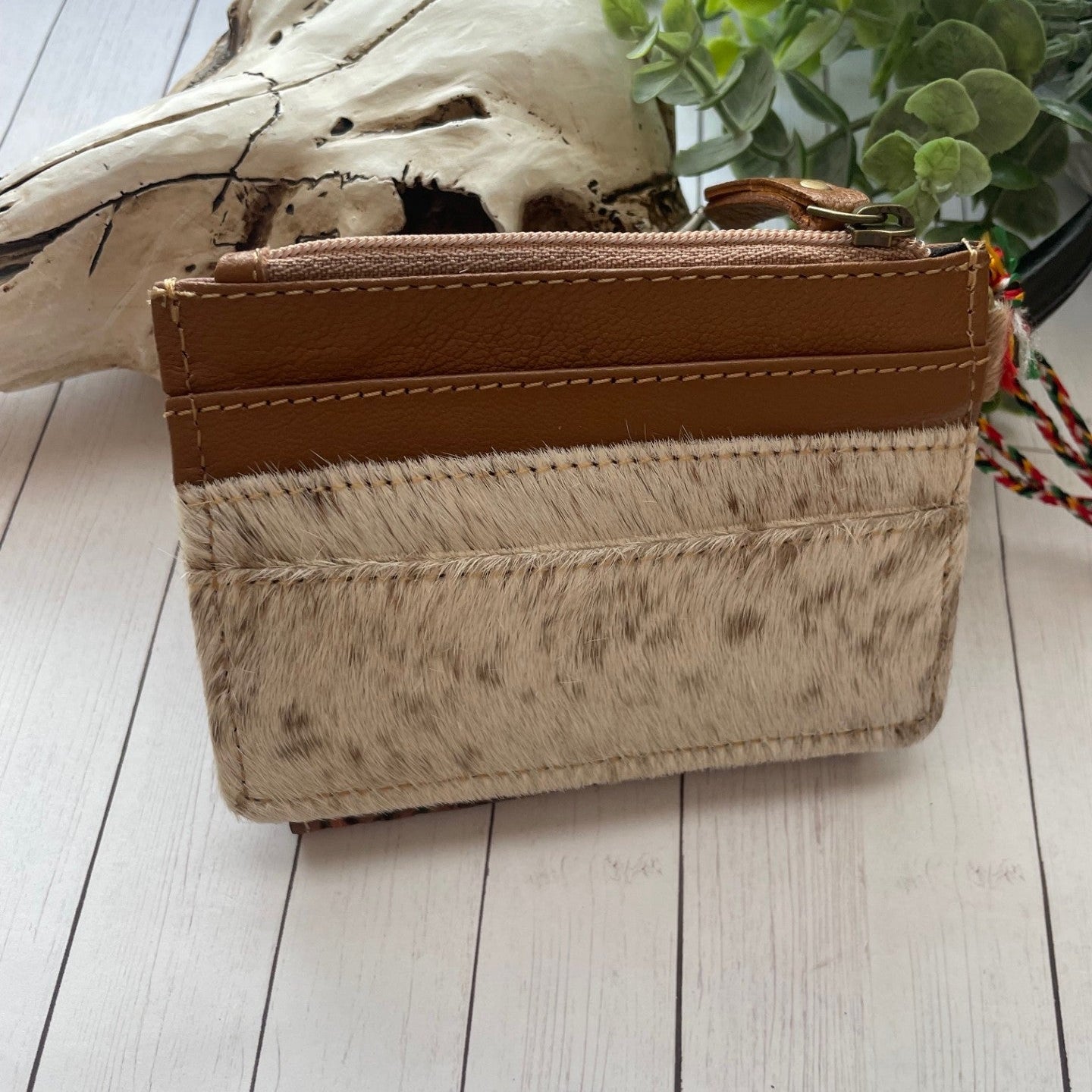 Brown leather wallet with brown and white cowhide and credit card slots on a wooden surface with plants in the background