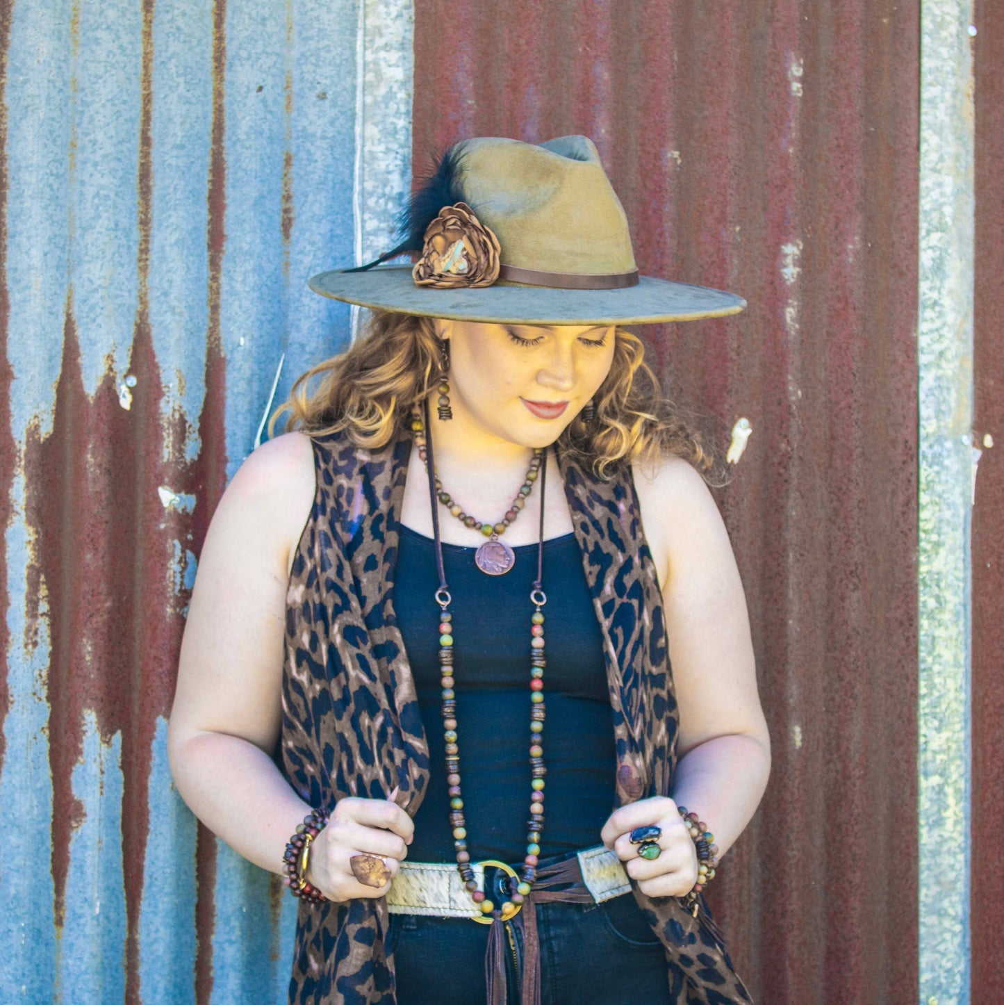 Woman wearing a hat with a feather and leopard print vest against a rustic corrugated metal wall.