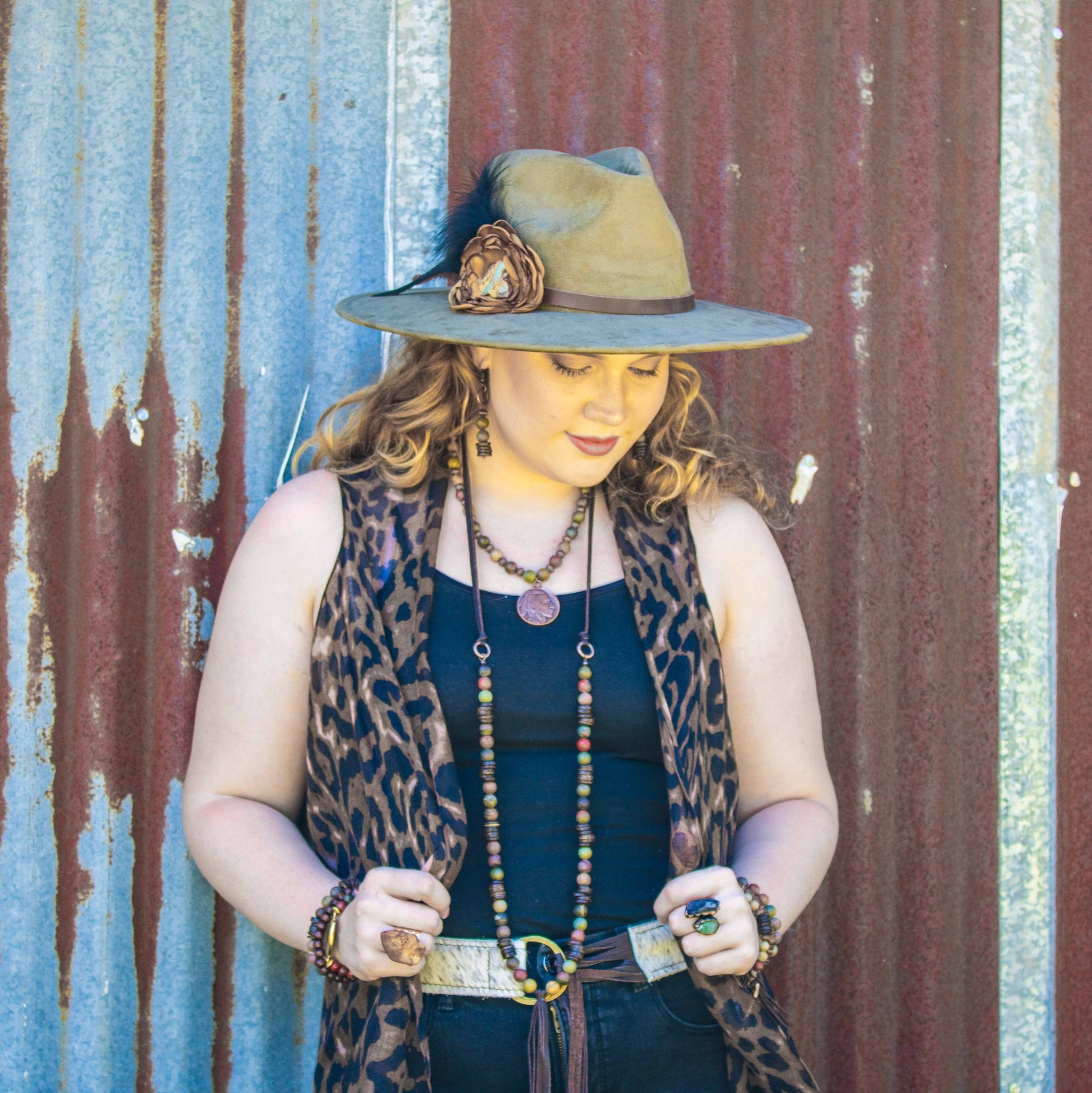 Woman wearing a hat with a feather and leopard print vest against a rustic corrugated metal wall.