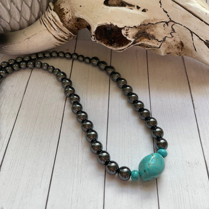 Necklace with turquoise bead and black beads on a wooden surface with a skull in the background