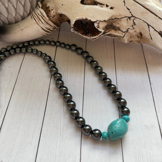 Necklace with turquoise bead and black beads on a wooden surface with a skull in the background