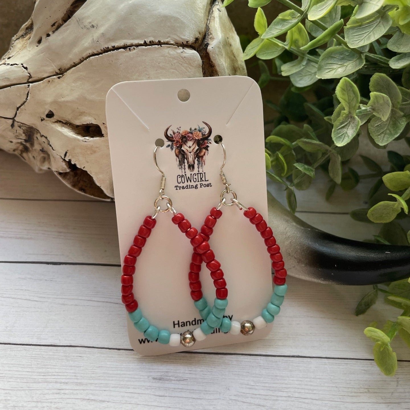 Red and turquoise beaded earrings on a white card with 'Cowgirl Trading Post' branding, placed on a wooden surface with driftwood and greenery.