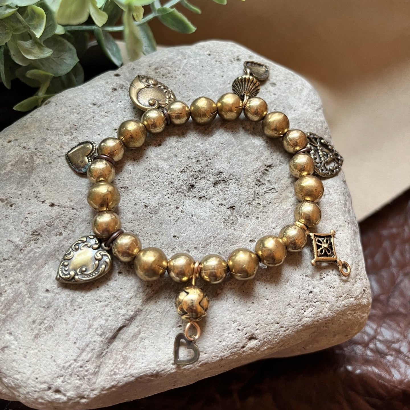 A gold-toned beaded charm bracelet displayed on a light gray stone, with decorative green leaves and a beige fabric background.
