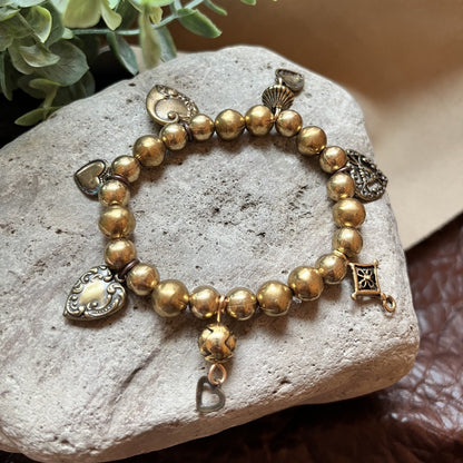 A gold-toned beaded charm bracelet displayed on a light gray stone, with decorative green leaves and a beige fabric background.