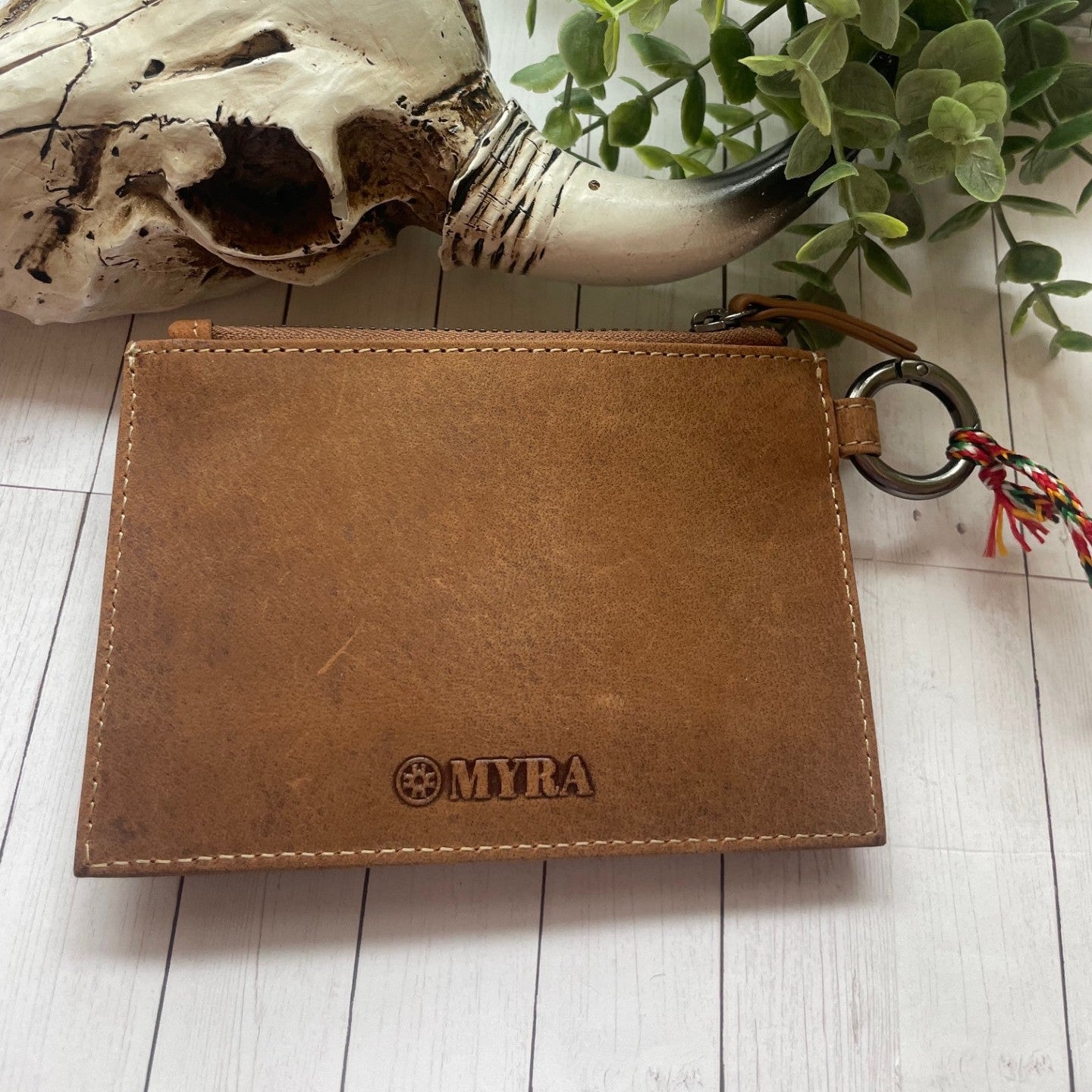 Brown leather wallet with 'MITRA' branding on a light wooden surface with a skull and plant in the background.