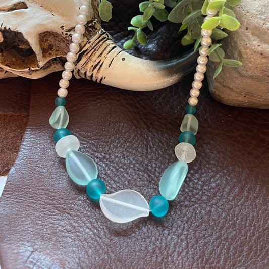 A beaded necklace with light blue and turquoise stones, white pearl-like beads, and translucent white teardrop beads, displayed on a brown leather surface with a skull, green plant, and stone props.