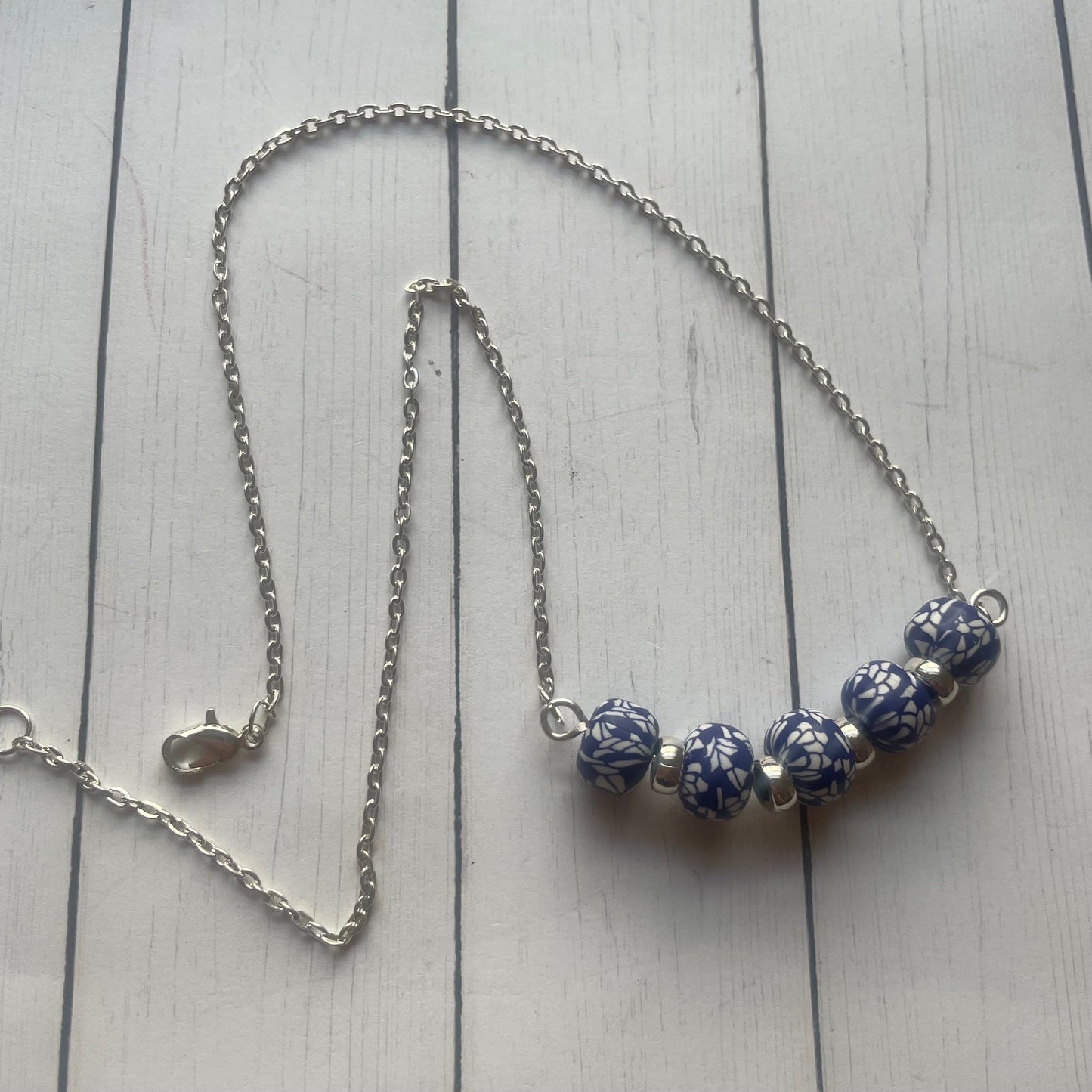 A silver chain necklace with five blue and white patterned beads, displayed on a light gray wooden plank background.