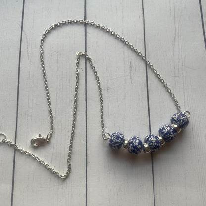 A silver chain necklace with five blue and white patterned beads, displayed on a light gray wooden plank background.