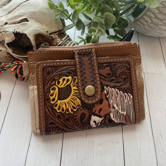 Brown leather wallet with the face head of a horse and a sunflower design on a wooden surface with plants in the background