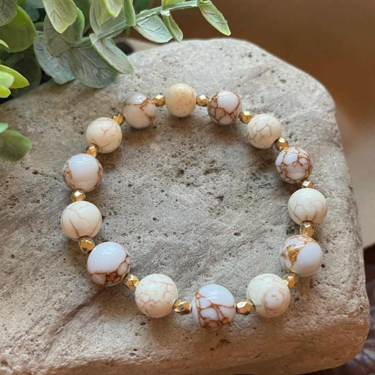 A beaded stretch bracelet made of white and cream marbled beads with brown veining, separated by small gold spacer beads, displayed on a textured stone with green leaves in the corner.