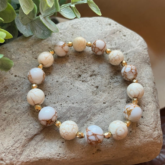 A beaded stretch bracelet made of white and cream marbled beads with brown veining, separated by small gold spacer beads, displayed on a textured stone with green leaves in the corner.