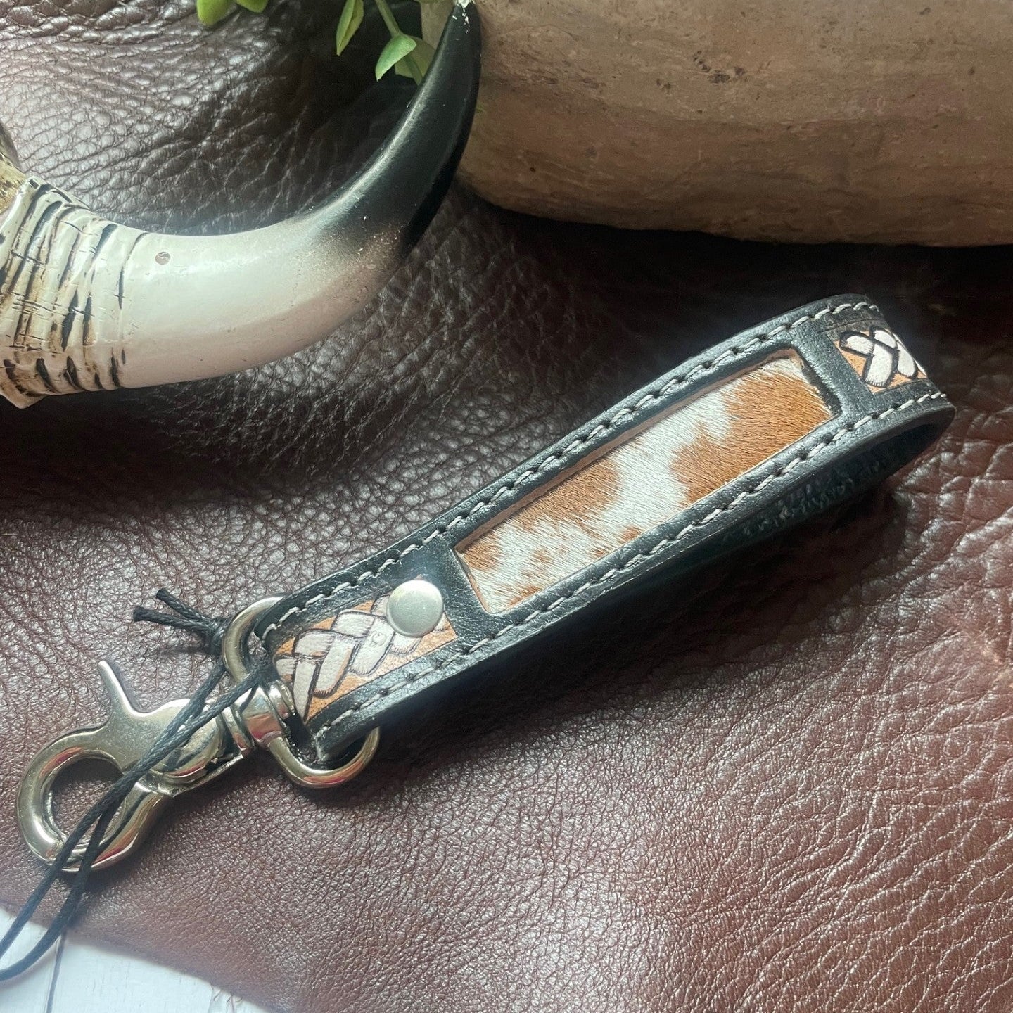 A close-up of a leather keychain or bag charm with a brown and white cowhide patch, stitched edges, braided trim, and a silver-tone metal clasp, resting on a brown leather surface with decorative props.