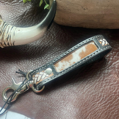 A close-up of a leather keychain or bag charm with a brown and white cowhide patch, stitched edges, braided trim, and a silver-tone metal clasp, resting on a brown leather surface with decorative props.