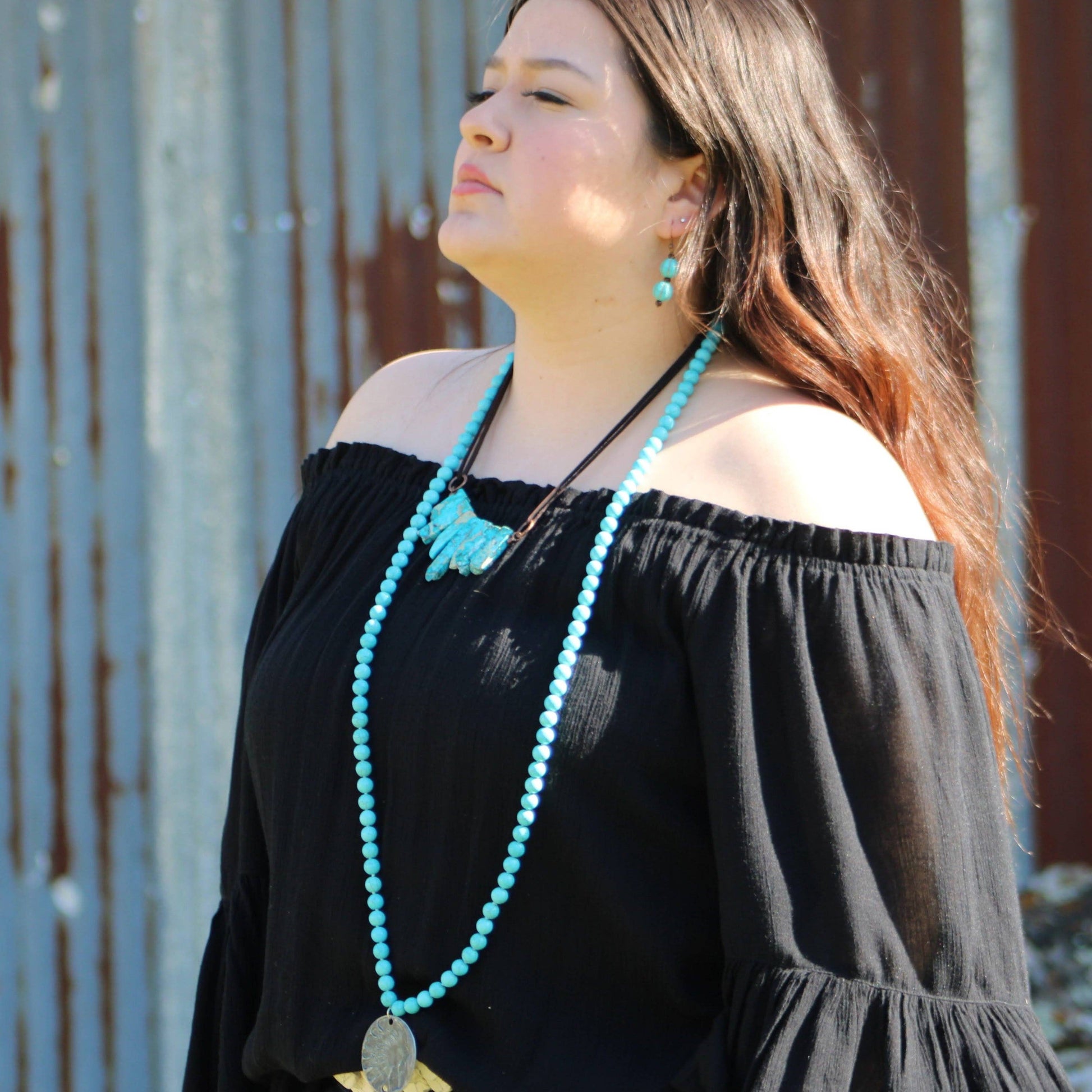 Woman wearing a black off-shoulder top with a long turquoise necklace against a rustic metal wall.