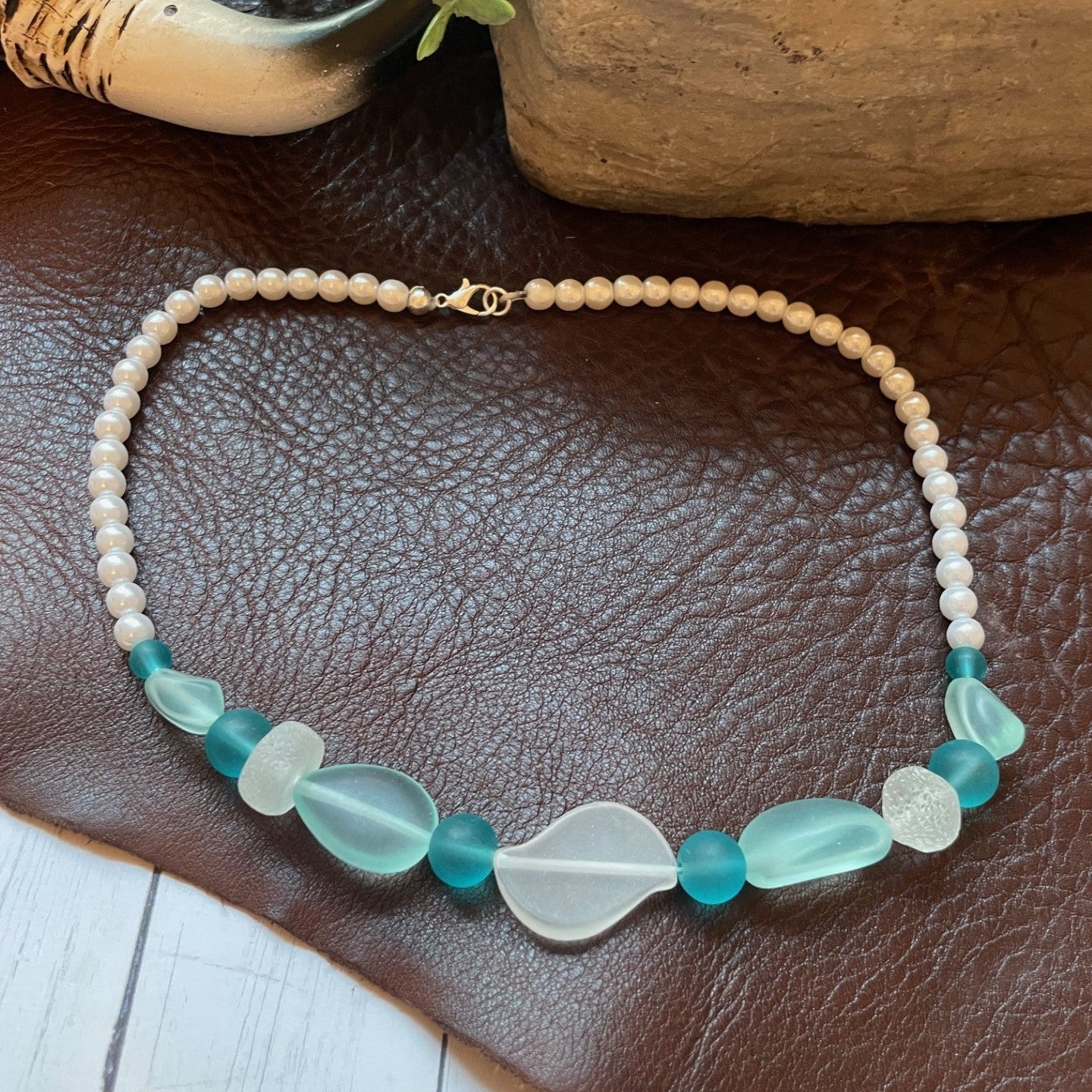 A beaded necklace with a strand of cream beads and a central section featuring translucent sea glass beads in teal and white, displayed on a brown leather surface.
