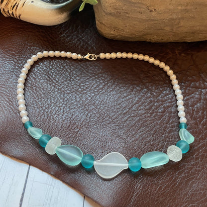 A beaded necklace with a strand of cream beads and a central section featuring translucent sea glass beads in teal and white, displayed on a brown leather surface.
